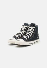Zapatillas de lona de caña alta en negro con cordones blancos, punteras de goma y detalles de costura prominentes. Tejido texturizado y suela contrastante.