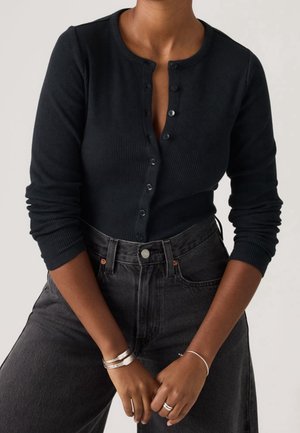Woman wearing a black ribbed long-sleeve buttoned top and high-waisted dark jeans, with silver bracelets and rings on her hands.