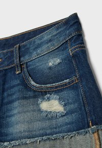 Shorts en denim bleu avec bords effilochés et empiècements usés près de la poche et sur la cuisse supérieure, montrant des détails de couture visibles.