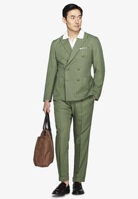 Mand iført lys grøn stribet dobbeltradet jakkesæt med hvid skjorte, holder en brun shopper taske og har sorte loafers på, stående mod en hvid baggrund.