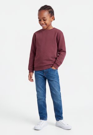 Barn iført en vinrød langærmet skjorte, blå jeans og hvide sneakers, står afslappet med den ene hånd i lommen og smiler nedad.