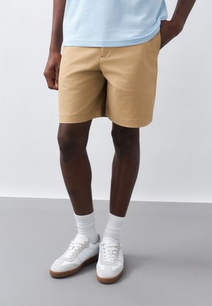 Άνδρας που φοράει μπεζ σορτς, γαλάζιο πουκάμισο, λευκές κάλτσες και λευκά αθλητικά παπούτσια, στέκεται σε γκρι πάτωμα με μονόχρωμο φόντο.