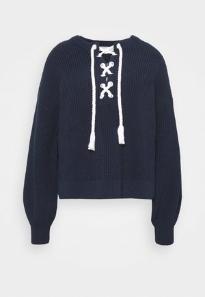 Maglione lavorato a maglia blu navy con un design cropped, scollatura rotonda e dettaglio bianco con lacci sul davanti che presenta dei pon-pon alle estremità.