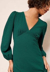 Rochie verde cu textură, cu decolteu adânc în V, mâneci lungi cu volane și design înfășurat. Materialul prezintă un model subtil, crăpat.
