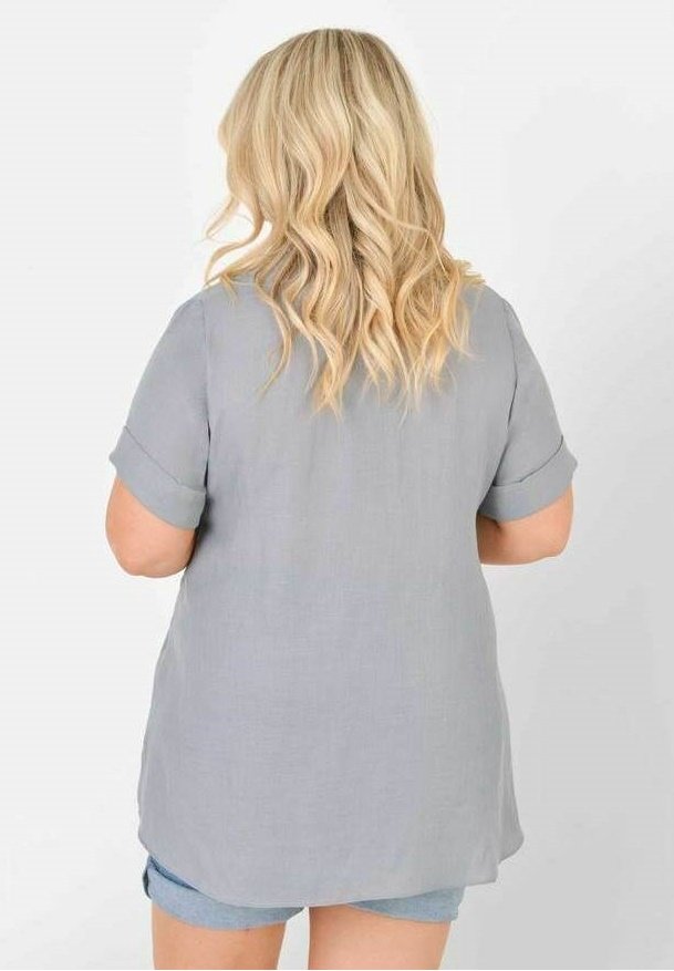 Světle šedá košile s krátkým rukávem a ohrnutými manžetami, která má volnější střih a delší zadní lem. Vyrobena z prodyšné tkaniny.
