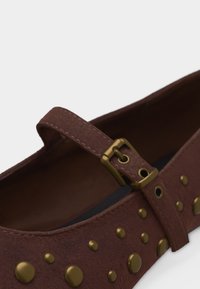 Chaussure plate en cuir marron avec une bride à boucle dorée et des clous ronds dorés décorant le dessus.