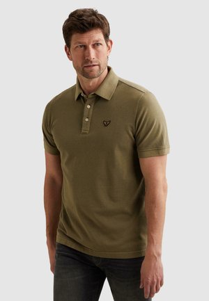 Man draagt een olijfgroene polo met korte mouwen en geborduurd logo, kijkt iets opzij tegen een effen achtergrond.