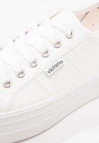 Baskets en toile blanches avec œillets ronds en argent, lacets blancs, semelle en caoutchouc épaisse et une petite étiquette de marque sur le côté.