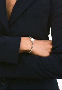 Braccialetto a catena d'argento con un grande accento di perla bianca, indossato su un polso nudo, su uno sfondo di blazer scuro. Design semplice ed elegante.