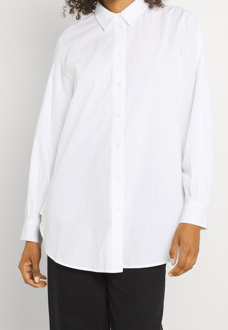 Chemise blanche boutonnée avec un col classique, manches longues et poignets élastiqués ; fabriquée dans un tissu lisse avec une coupe droite et ample.
