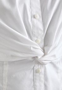 Bijela košulja s dugmadima, s čvorom sprijeda, s glatkom teksturom, zaobljenim ovratnikom i šest bijelih dugmadi duž plisa.