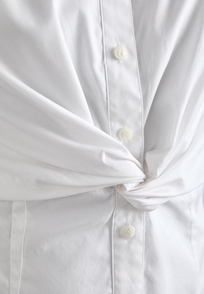 Camicia bianca con un design annodato in vita, realizzata in tessuto morbido, con cinque bottoni e dettagli di cucitura discreti.