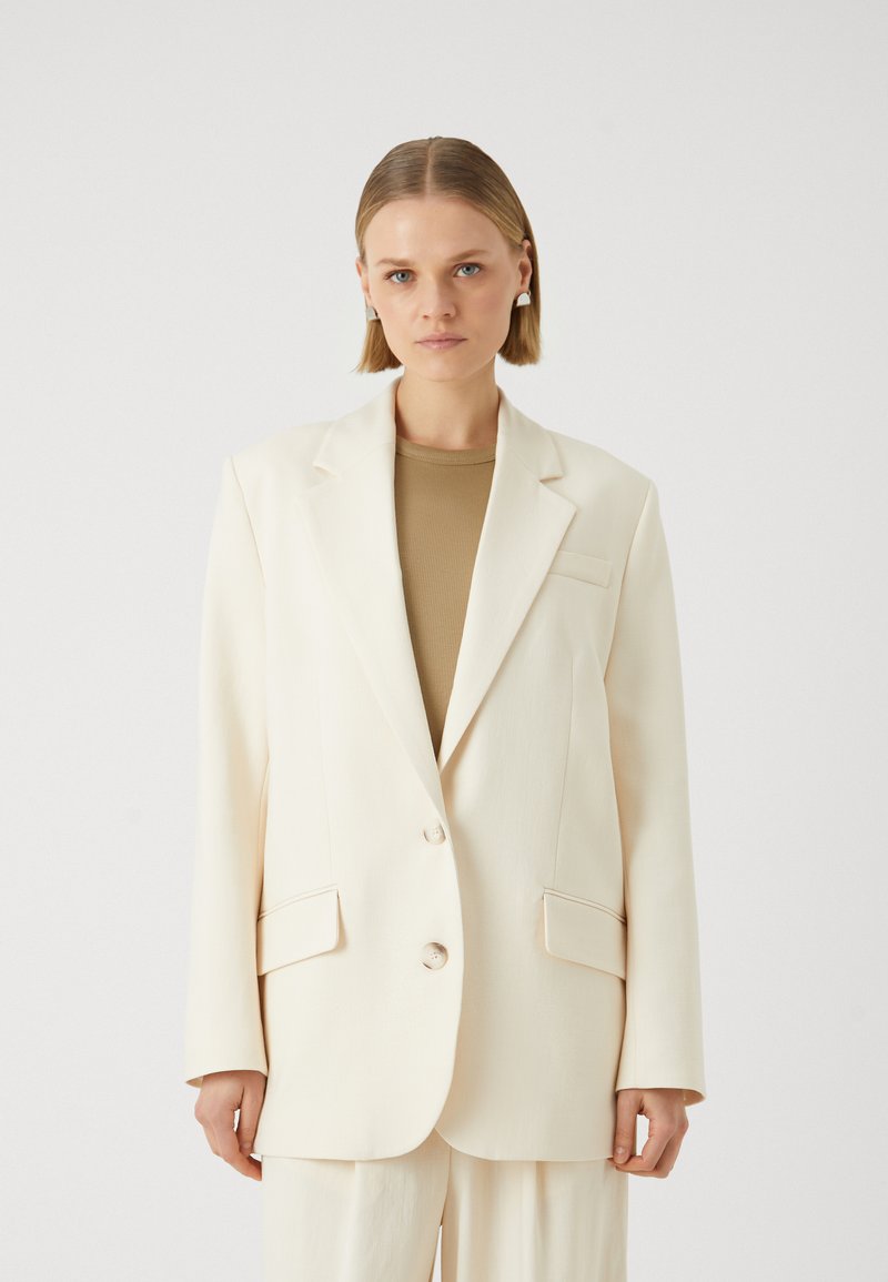 Blazer color crema con due bottoni, rever a punta e tasche frontali. Vestibilità sartoriale con una texture liscia, indossato sopra un top beige.
