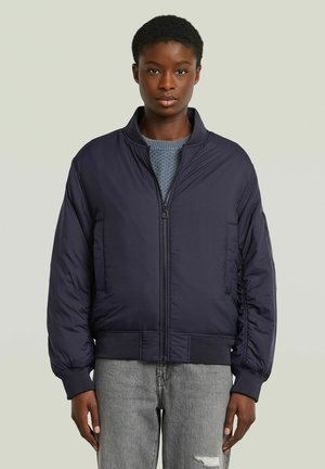 Giacca bomber blu navy con zip frontale, polsini e orlo a coste, e tasche laterali. Indossata sopra un top lavorato a maglia blu chiaro con un motivo testurizzato.