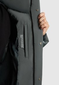 Sötétzöld párnázott dzseki, cipzáros záródással, patenttal, belső zsebbel és care utasításokat tartalmazó textil címkével.
