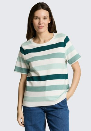 Kobieta z długimi ciemnymi włosami, ubrana w zielono-biały pasiasty krótki rękaw i niebieskie dżinsy, stoi z jedną ręką w kieszeni.