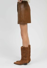 Femme portant une jupe en cuir brun avec des clous et des bottes de cow-boy en daim brodé marron, se tenant devant un fond clair.