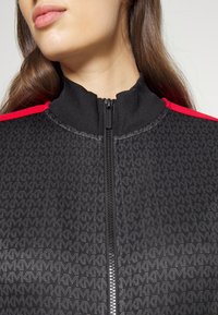 Svart zip-up jacka med hög krage, med röda axeldetaljer och ett mönstrat textur. Dragkedjan går vertikalt ner i mitten.