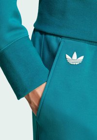 Teal träningströja med ribbade ärmar, en framficka och en broderad vit logotyp på sidan. Slät textur och figursydd design.