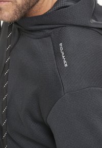 Mustat hupparit, joissa on teksturoitu kangas. Sisältää ommellun "ENDURANCE"-logon olkapäällä ja kuvioidun nauhan, joka parantaa muotoilua.