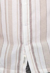 Chemise à rayures avec des couleurs blanche et marron clair, tissu texturé et boutons en plastique transparent. Étiquette de marque visible à l'ourlet.