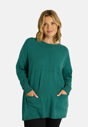 Femme aux cheveux blonds mi-longs, portant un pull vert ample à manches longues avec des poches et un pantalon noir, sur un fond uni.