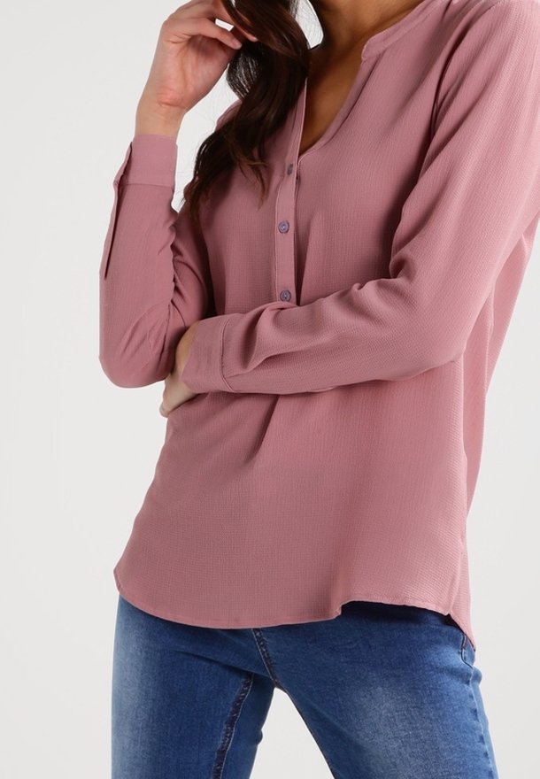 Femme portant un chemisier boutonné rose poudré à manches longues, associé à un jean bleu, posant avec une main près du visage et l’autre croisée.