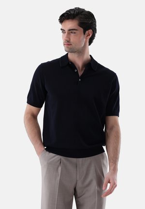 Mann mit dunklen Haaren, der ein schwarzes kurzärmeliges Poloshirt und beige Hose trägt, steht mit einer Hand in der Tasche vor einem einfarbigen Hintergrund.
