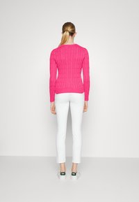 Suéter de punto en color rosa con puños y dobladillo acanalados, combinado con pantalones ajustados blancos y zapatillas blancas con acentos verdes, visto desde atrás.