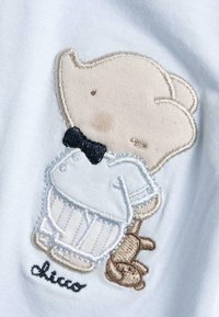 Personaggio ricamato con caratteristiche chiare, vestito di bianco con un papillon nero, tiene in mano un orsetto di peluche marrone, su uno sfondo blu chiaro.