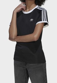 Svart atletisk t-shirt med vit krage och ärmdekorer, med tre vita ränder på varje axel och en liten Adidas-logga på bröstet.