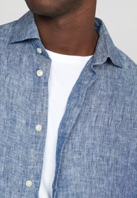 Camicia di lino azzurro chiaro con una finitura testurizzata, colletto button-down e una maglietta bianca sotto. Presenta bottoni in perla e dettagli di cucitura visibili.