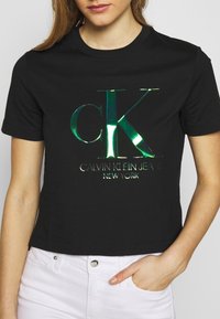 Černé zkrácené tričko s lesklým zeleným logem "CK" a textem "CALVIN KLEIN JEANS NEW YORK" pod ním, vyrobené z jemné bavlny.