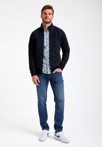 Veste zippée matelassée bleu marine sur une chemise à motifs, assortie à un jean bleu et des sneakers blanches avec des accents noirs.