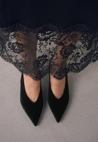 Pantofi cu toc ascuțit din catifea neagră, plasați deasupra unei margini din dantelă neagră cu modele florale și o margine franjurată, pe un fundal neutru.