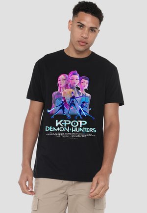 Young man wearing black "K-Pop Demon Hunters" t-shirt featuring animated female characters with glowing weapons and neon text.