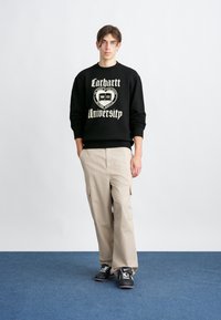 Czarna bluza z grafiką "Carhartt University", beżowe spodnie cargo i czarne sneakersy, stojący na niebieskim dywanie na białym tle.