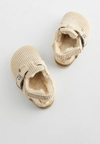 Beige kordflöjtslippers med en mjuk pälsfoder, justerbar rem med silverfärgade spännen och platt sula, som visar en ribbad textur.