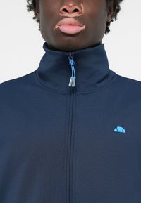Mornarska zip-up jakna z visokim ovratnikom, izdelana iz gladkega materiala. Ima svetlo modro padajočo logotip in zadržne detajle. Prikazan je podroben pogled.