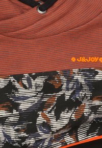 Multicolor hoodie met een textuur in roestkleur aan de bovenkant en een grafisch zwart-wit bloemenpatroon eronder. Beschikt over een trekkoord en een oranje logo.