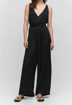 Kobieta ubrana w czarny, szeroki kombinezon na ramiączkach, z kopertowym przodem i wiązaniem w talii, w połączeniu z czarnymi, płaskimi sandałami z ćwiekami.