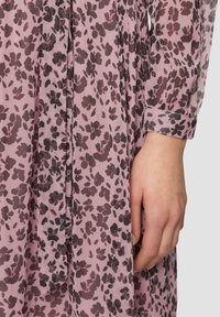 Rosa Kleid mit floralem Muster in Dunkelbraun. Hat lange Ärmel, eine gebundene Taille und einen fließenden, strukturierten Stoff. Nahaufnahme einer Hand auf dem Stoff.