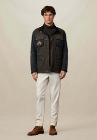 Mlad muškarac stoji u tamnozelenoj i mornarsko plavoj jakni, maslinastoj vratnoj dukserici, bijelim hlačama i smeškim suknjenim cipelama na bež pozadini.