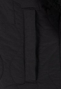 Schwarzer gesteppter Stoff mit strukturierter Oberfläche. Verfügt über eine vertikale Tasche mit einer sauberen Naht und minimalem Nähdetails.