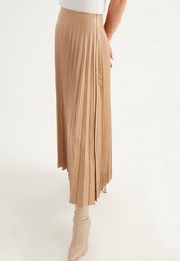 Plisserad midikjol i tan konstläder, med dragkedjestängning, slät yta och ett flödande snitt som når ner till anklarna.