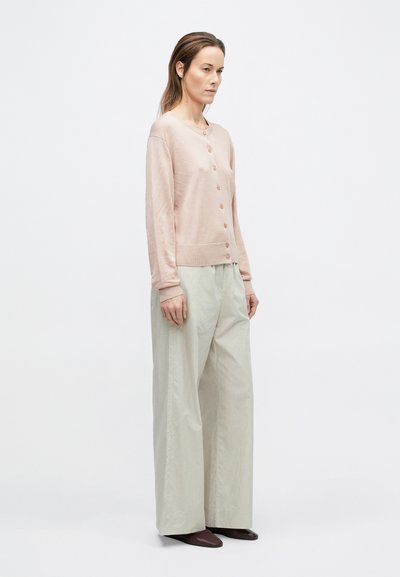 Frau trägt hellrosa geknöpften Cardigan, weit geschnittene hellbeige Hose und dunkle spitze Schuhe, steht vor einfachem hellem Hintergrund.