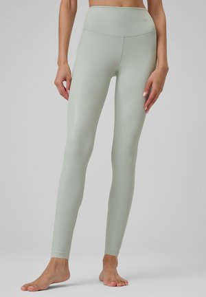 Legginsy slim fit w jasnym zielonym kolorze, z wysokim stanem, noszone boso, skrócony środkowy fragment z rękami luźno opartymi po bokach, jednolite szare tło.