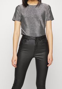 Person som bär en silverglittrig kortärmad topp nedstoppad i högmidjade svarta läderliknande byxor, stående mot en enkel bakgrund.