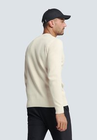Crème kleurige geribbelde trui met lange mouwen, ronde hals en een aansluitende zoom, gecombineerd met een zwarte pet. Model wordt vanuit een zijaanzicht getoond.