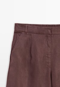 Gros plan sur un pantalon marron montrant la ceinture, les passants de ceinture, la poche latérale et les plis avant.